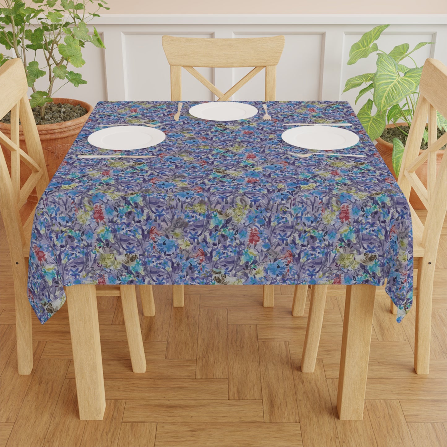 Dining table set with a floral tablecloth on a wooden floor. Round table covered with a blue watercolor floral tablecloth featuring botanical patterns in blue, lavender, and green tones.