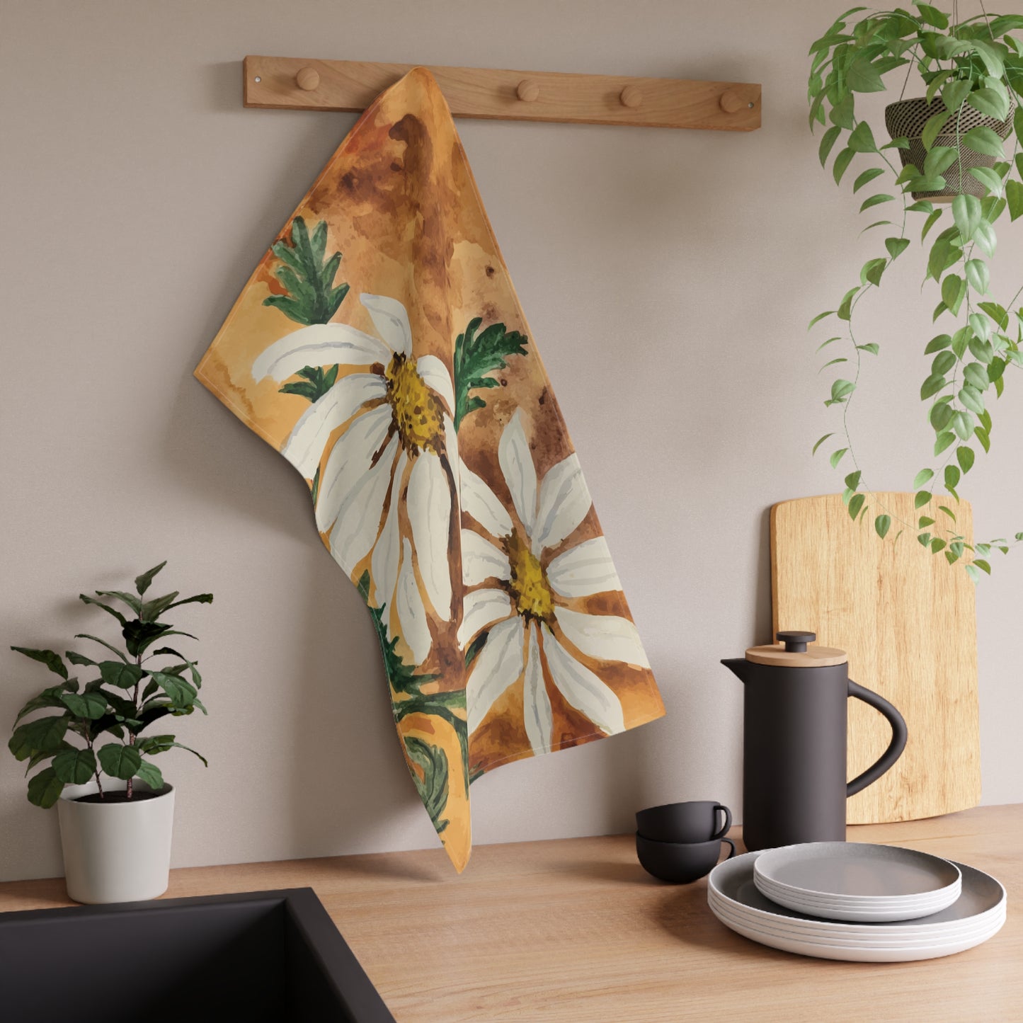 Watercolor daisy tea towel with white petals and golden centers on a warm orange background, displayed hanging in a bright kitchen. Floral towel hanging on a wooden rack with kitchen items and plants in the background