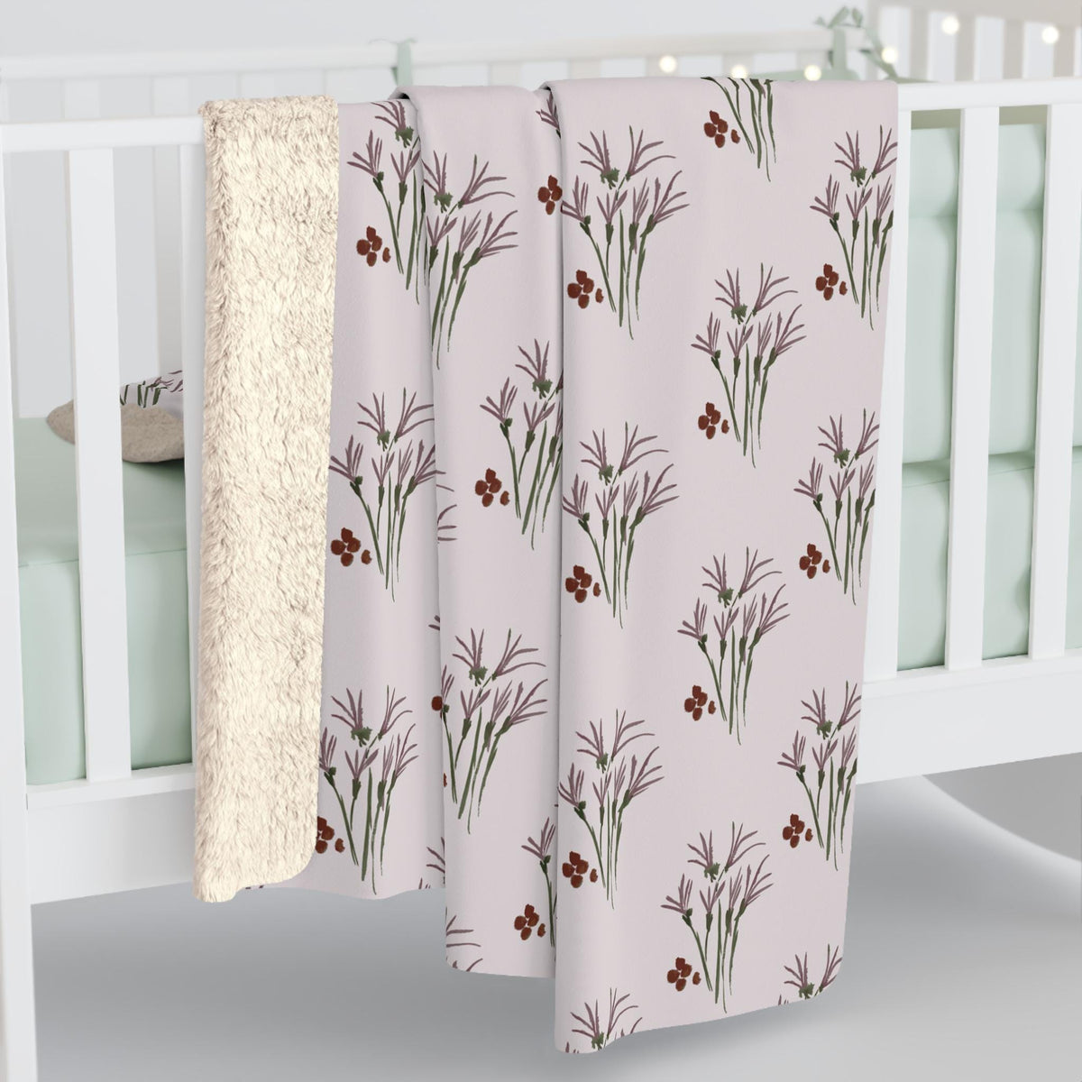 A light pink blanket with a repeating pattern of stylized purple flowers and red berries. The blanket has a soft, plush cream-colored backing. The blanket is draped over a white crib.