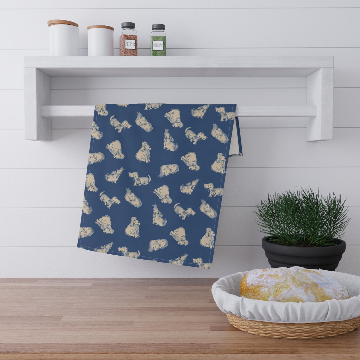 Navy blue kitchen tea towel featuring a repeating hand-drawn dog pattern in soft cream tones, displayed hanging in a bright kitchen with a wooden countertop, herbs, and fresh bread for cozy pet lover home décor.