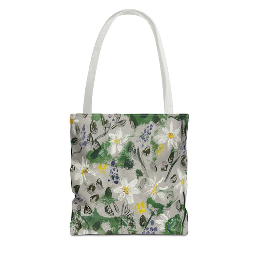 A canvas like  tote bag with delicate white wildflowers, soft lavender blossoms, leafy greens, and painterly botanical textures come together to create a timeless design inspired by peaceful forest walks and secret gardens waiting to be discovered, on a light gray background, complete with two white shoulder straps.