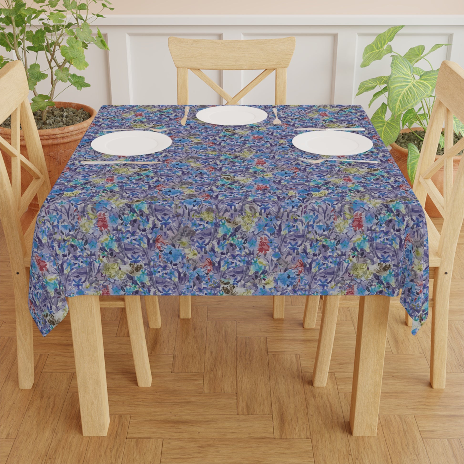 Dining table set with a floral tablecloth on a wooden floor. Round table covered with a blue watercolor floral tablecloth featuring botanical patterns in blue, lavender, and green tones.