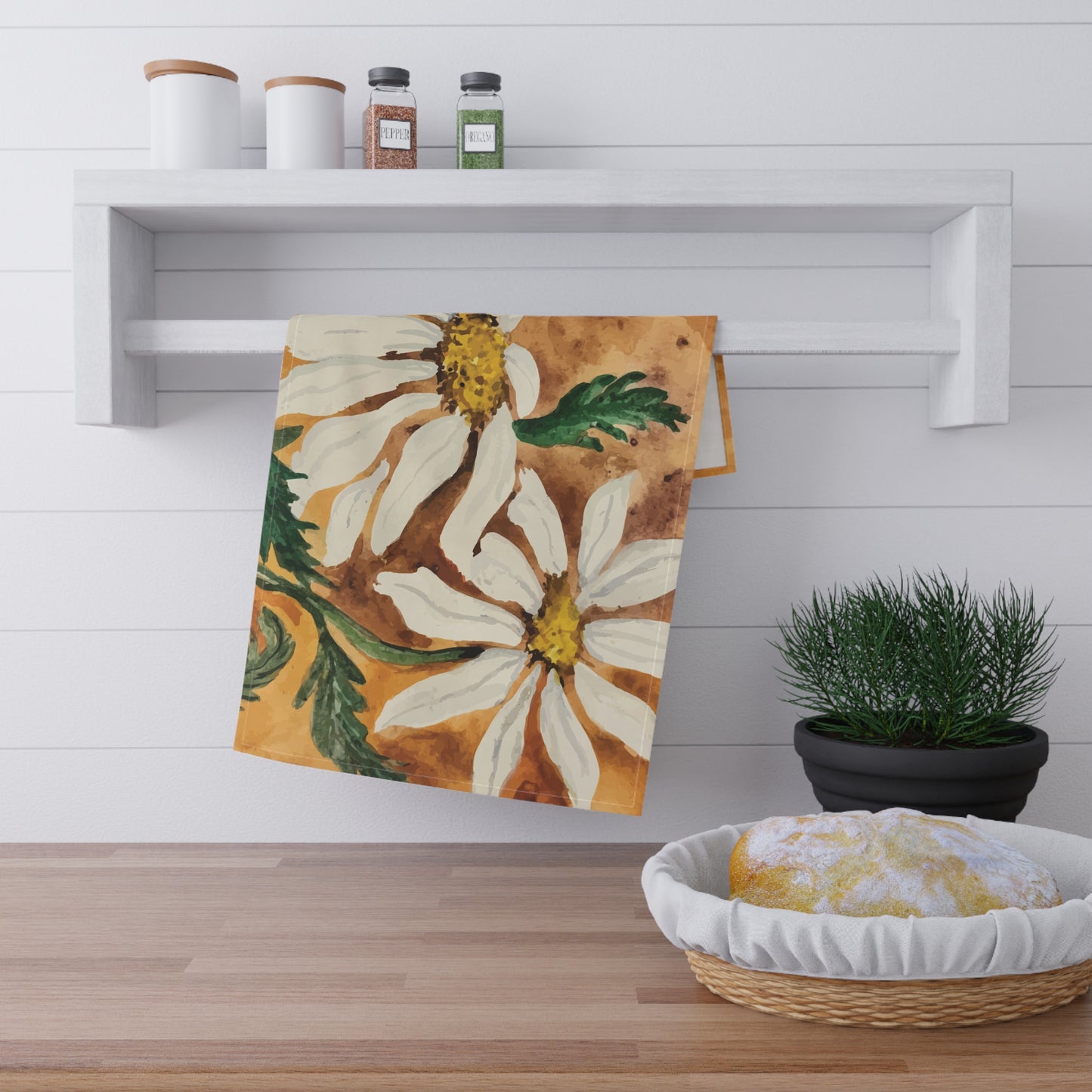 Watercolor daisy tea towel with white petals and golden centers on a warm orange background, displayed hanging in a bright kitchen.