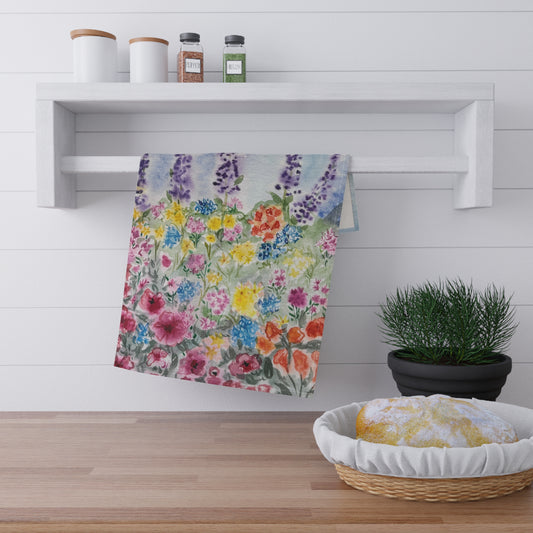 Watercolor floral tea towel featuring lavender stems and colorful wildflowers in soft blue, purple, pink, and yellow tones on a light background. Floral towel hanging on a rack with a kitchen setting including a basket and potted plant.