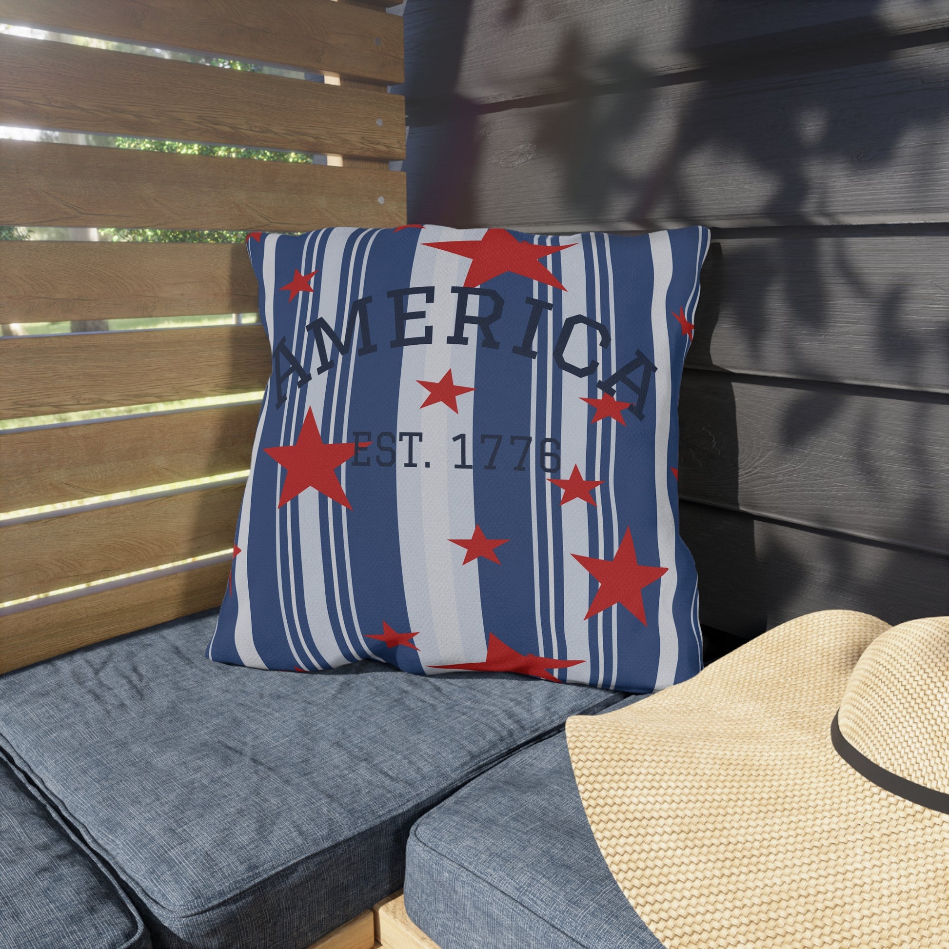 Decorative pillow with 'America Est. 1776' text and red stars on a blue background, placed on a wooden bench.