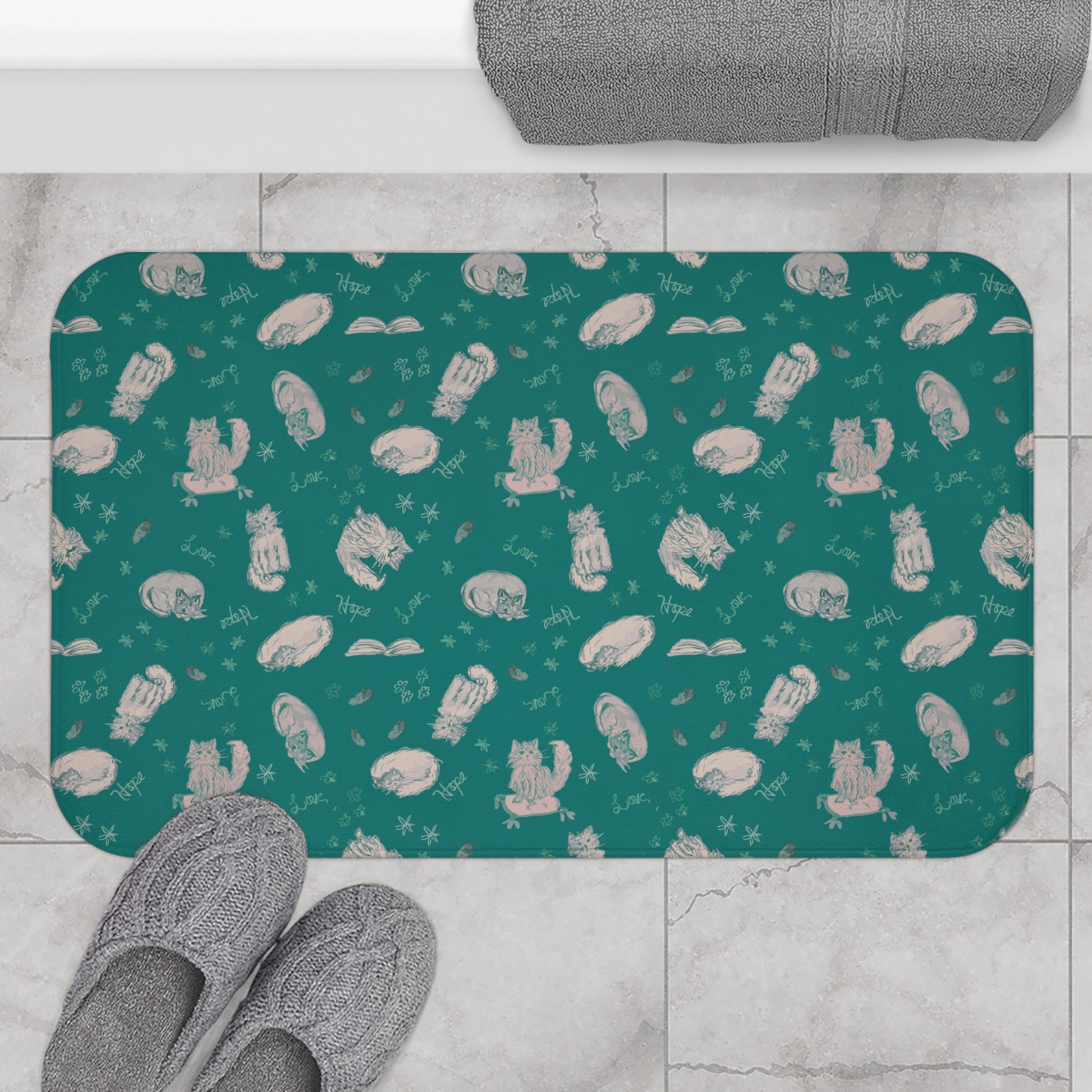 Teal bath mat with white cat pattern on a tiled floor with slippers.