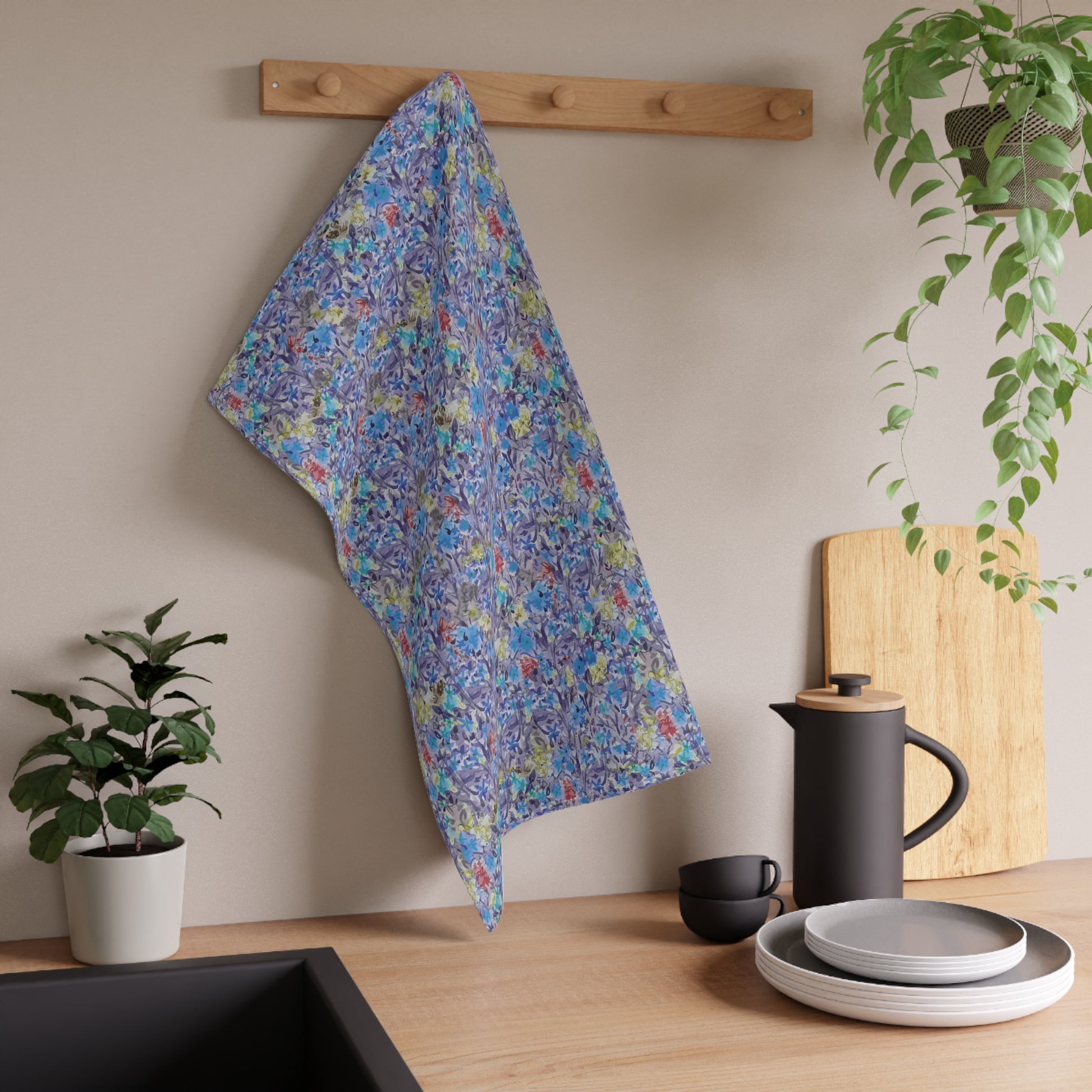 Watercolor floral tea towel with pink blossoms, earthy brown tones, and soft blue accents in a meadow-style pattern. Floral towel hanging on a wooden rack above a kitchen counter with plates, cups, and a plant.