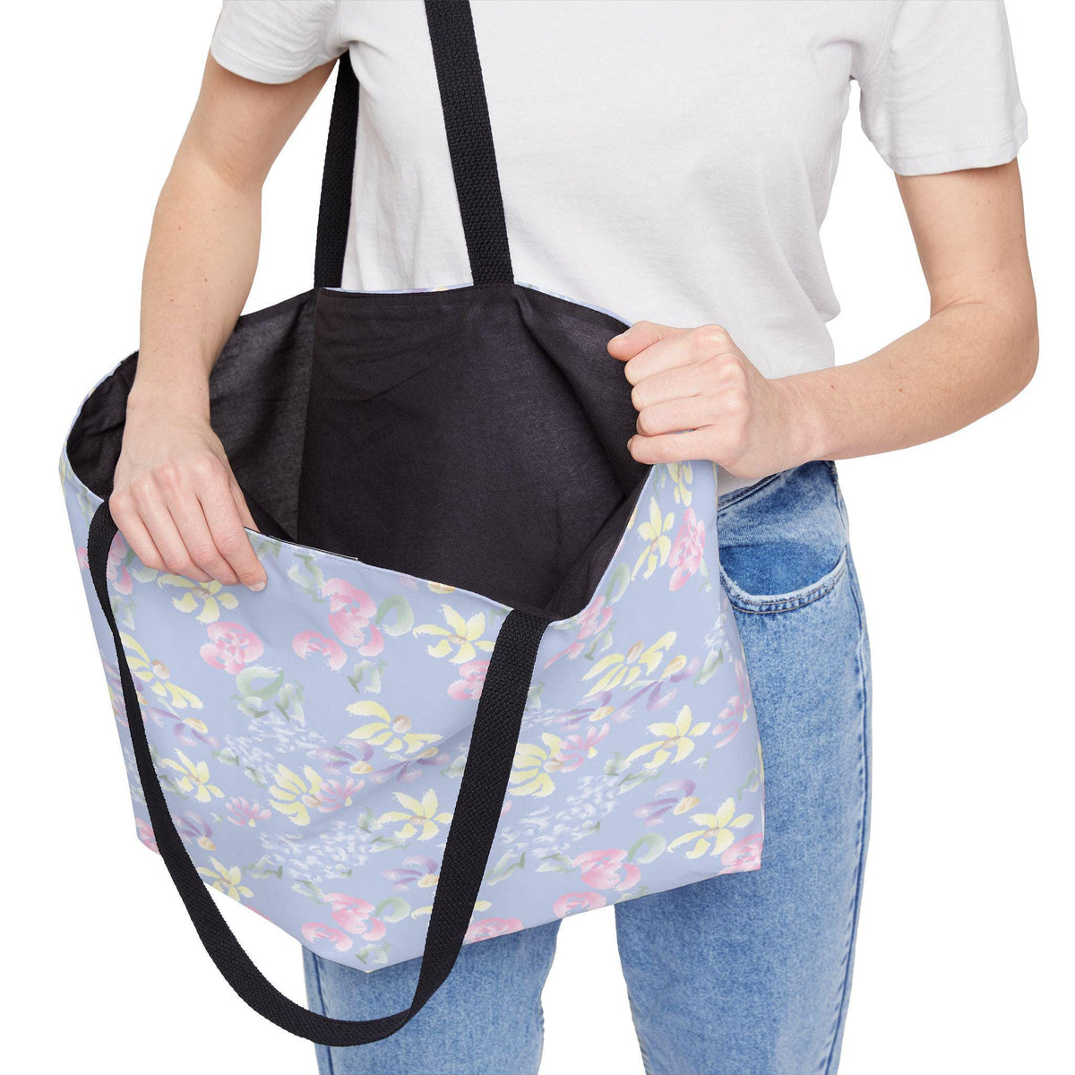 A light blue tote bag with a floral pattern in pink, yellow, and green. The bag has black straps and a black interior. The bag is being held open, revealing the interior.
