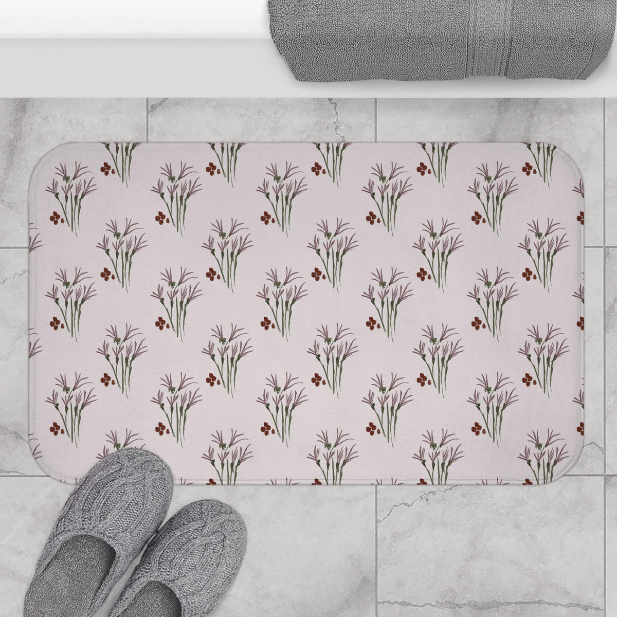 A rectangular bath mat with rounded corners, featuring a repeating floral pattern. The design includes purple flower clusters and small red berries on a light pink background. Gray slippers are visible at the bottom.