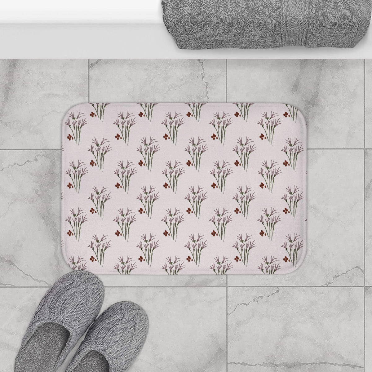 A rectangular bath mat with a repeating floral pattern. The design features stylized purple flowers with green stems and small red accents on a light pink background. Gray slippers are at the bottom.