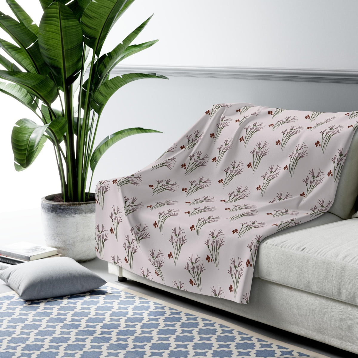 A light pink throw blanket with a repeating pattern of stylized floral sprigs in shades of brown and green. The blanket is draped over a white sofa. A large potted plant is visible in the background.