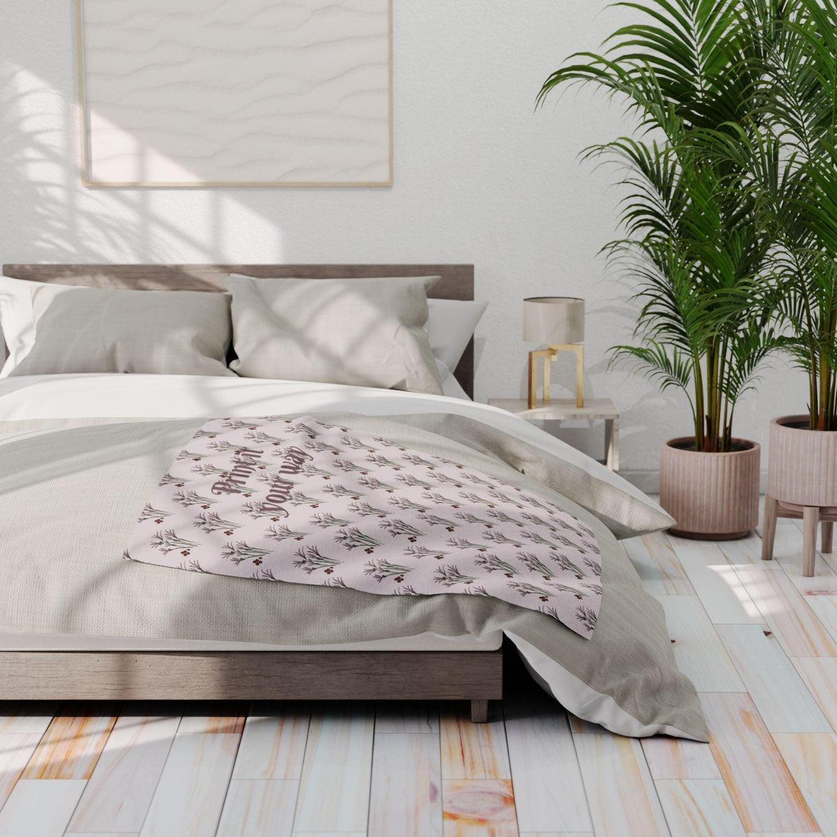 A light pink blanket with a repeating floral pattern and the text "Print it your way" draped over a bed. The bed has a wooden frame and is covered with light-colored bedding. A large framed artwork hangs on the wall.