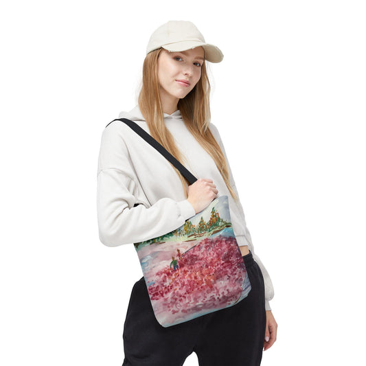 A white tote bag with a black shoulder strap featuring a watercolor painting of a landscape with red and pink flowers, green trees, and a blue sky. The bag is carried by a person wearing a white hoodie and a cream-colored baseball cap.