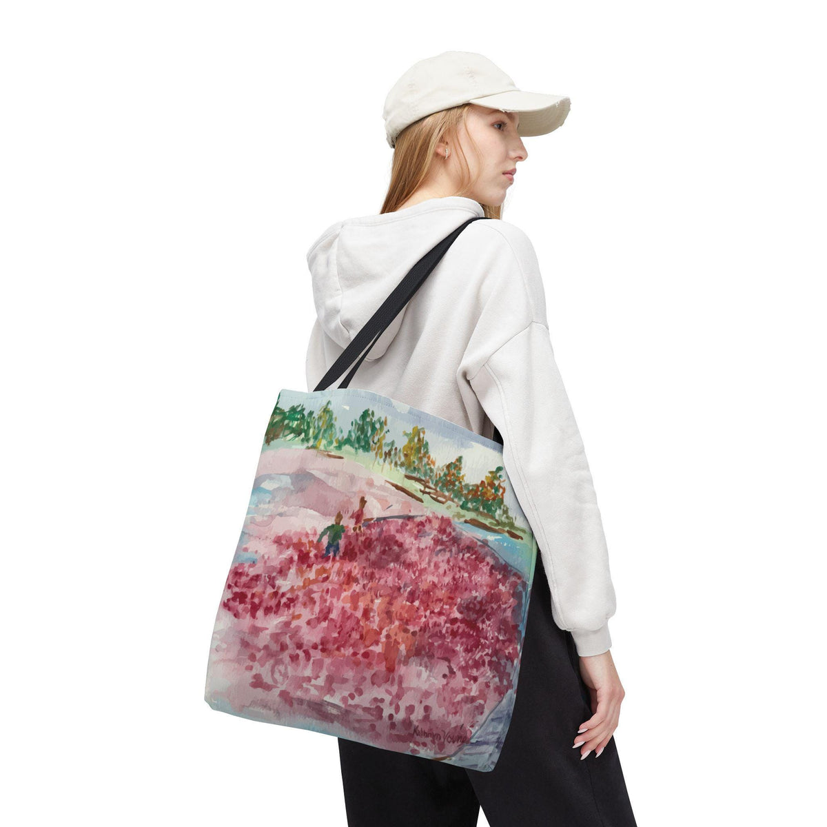 A tote bag with a watercolor painting of a landscape featuring a field of red flowers, green trees, and a blue sky. The bag has a black strap and is carried over the shoulder.