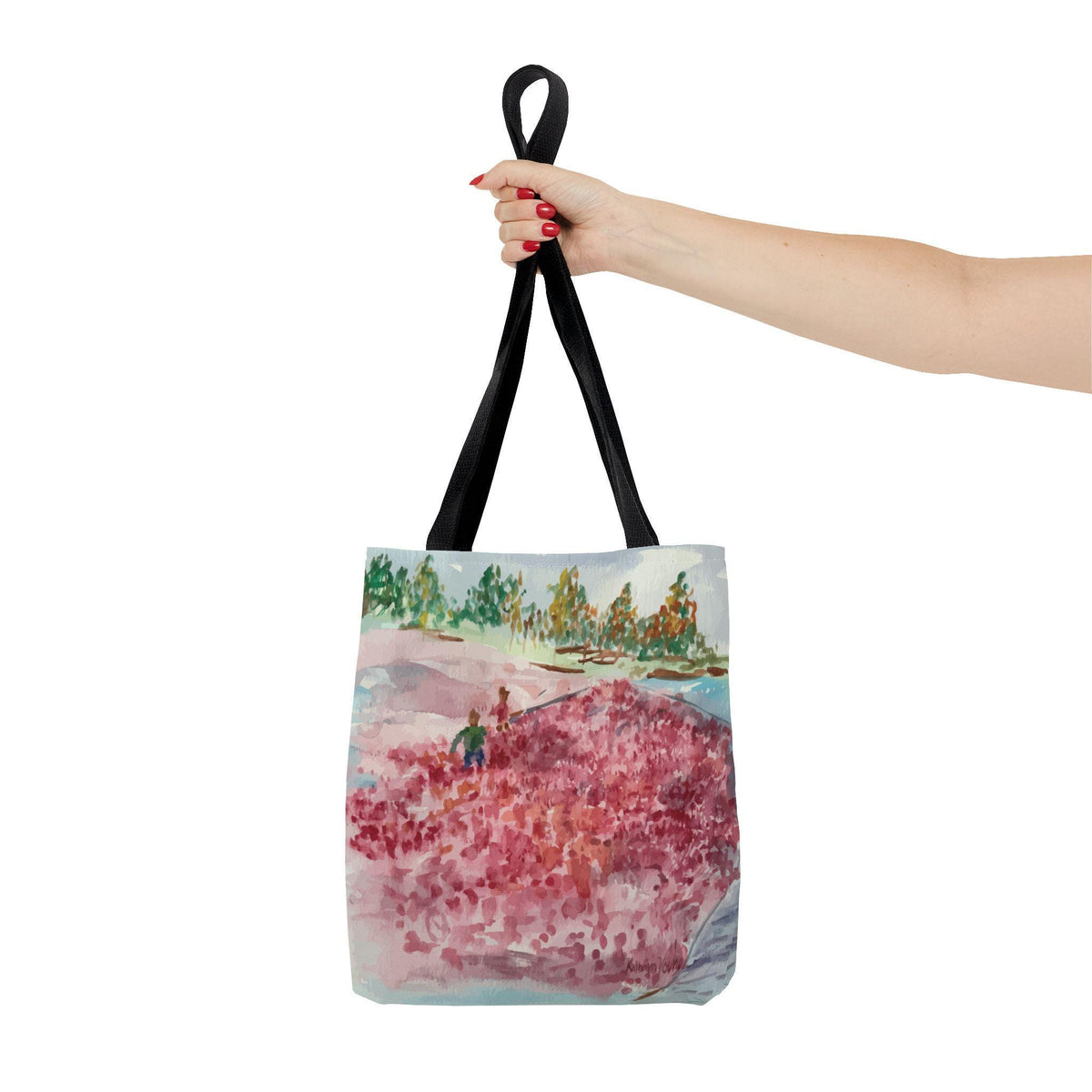 A tote bag with a watercolor painting of a landscape featuring red and pink flowers, green trees, and a blue sky. The bag has black handles and is held up by a hand.