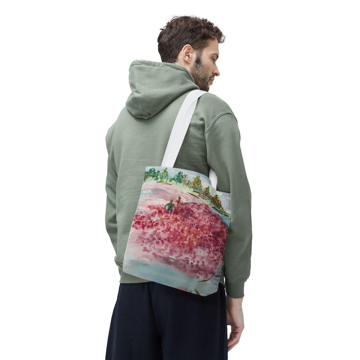 A white tote bag with a watercolor painting of a pink field, green trees, and two figures. The bag has a white strap and is carried over the shoulder. The person in the image is wearing a green hoodie and dark blue pants.