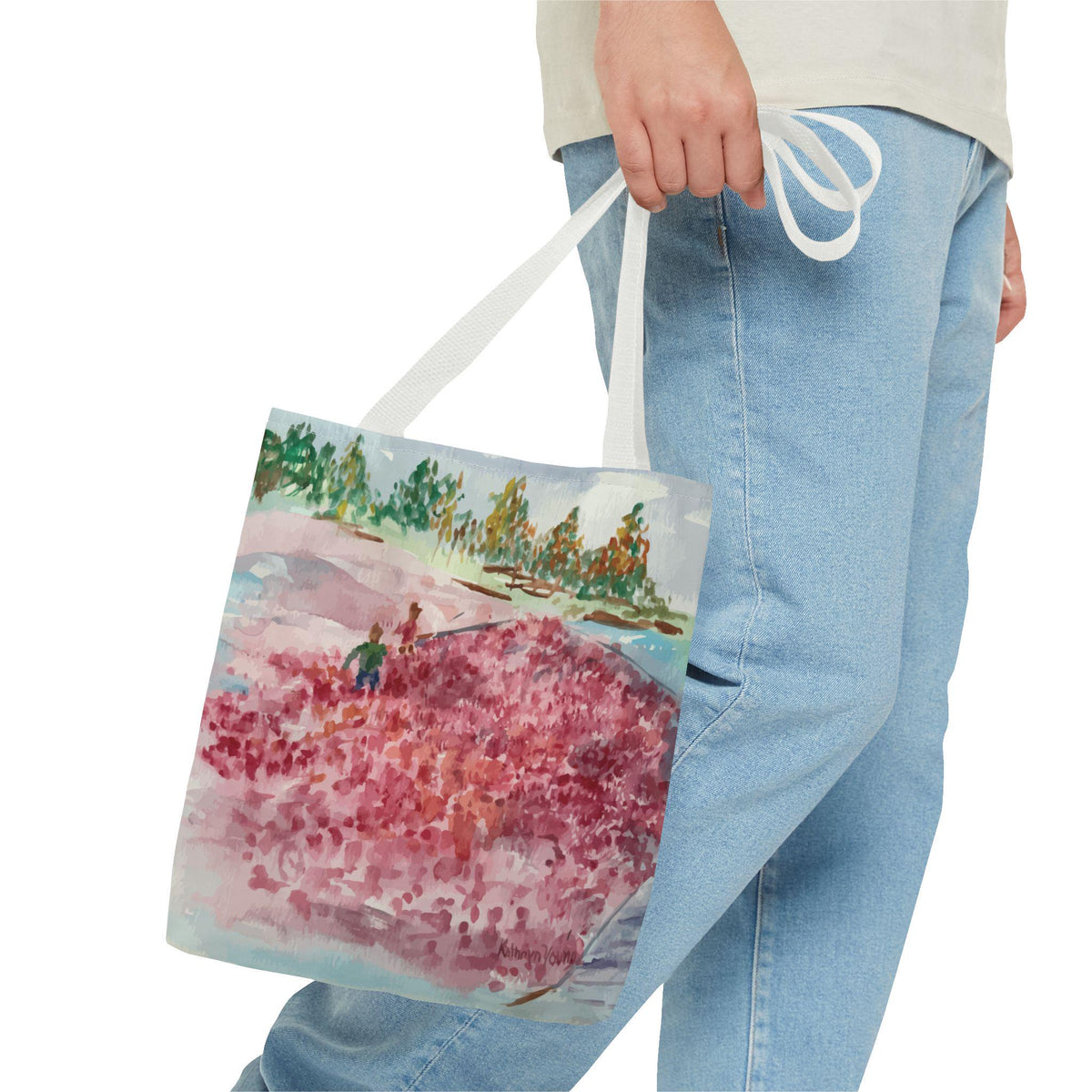A white tote bag featuring a watercolor painting of a cranberry bog. The artwork depicts a person in a green shirt standing in a field of red berries, with trees and a blue sky in the background. The bag has white straps.