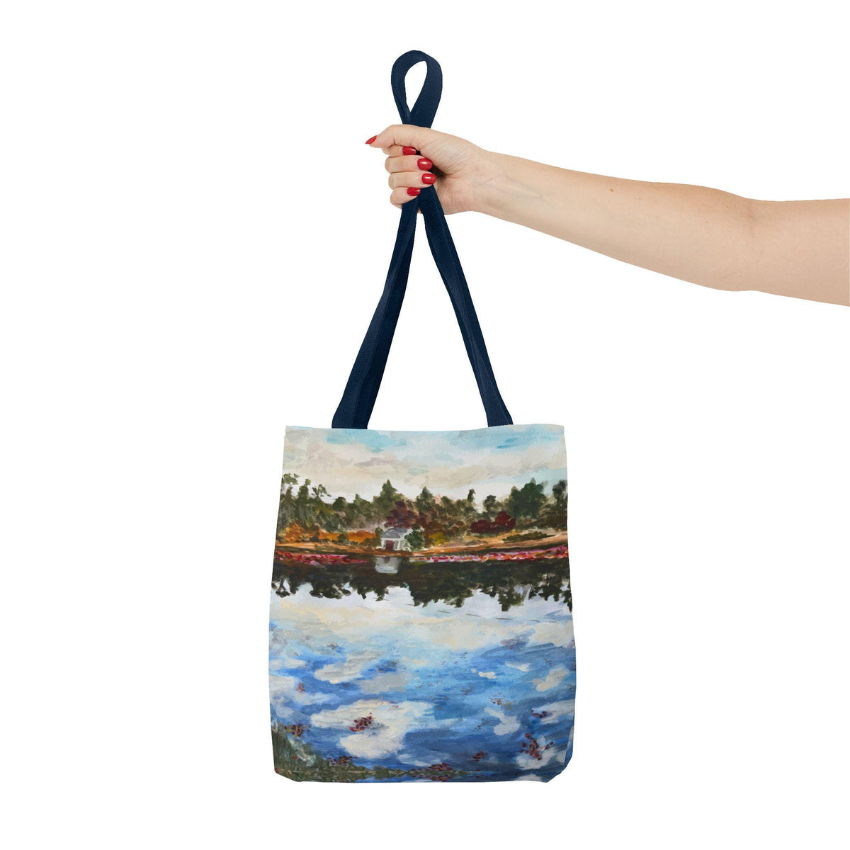A tote bag with a painting of a lake scene. The bag has a navy blue handle and a watercolor painting of a lake with trees, clouds, and reflections. The bag is held by a person.
