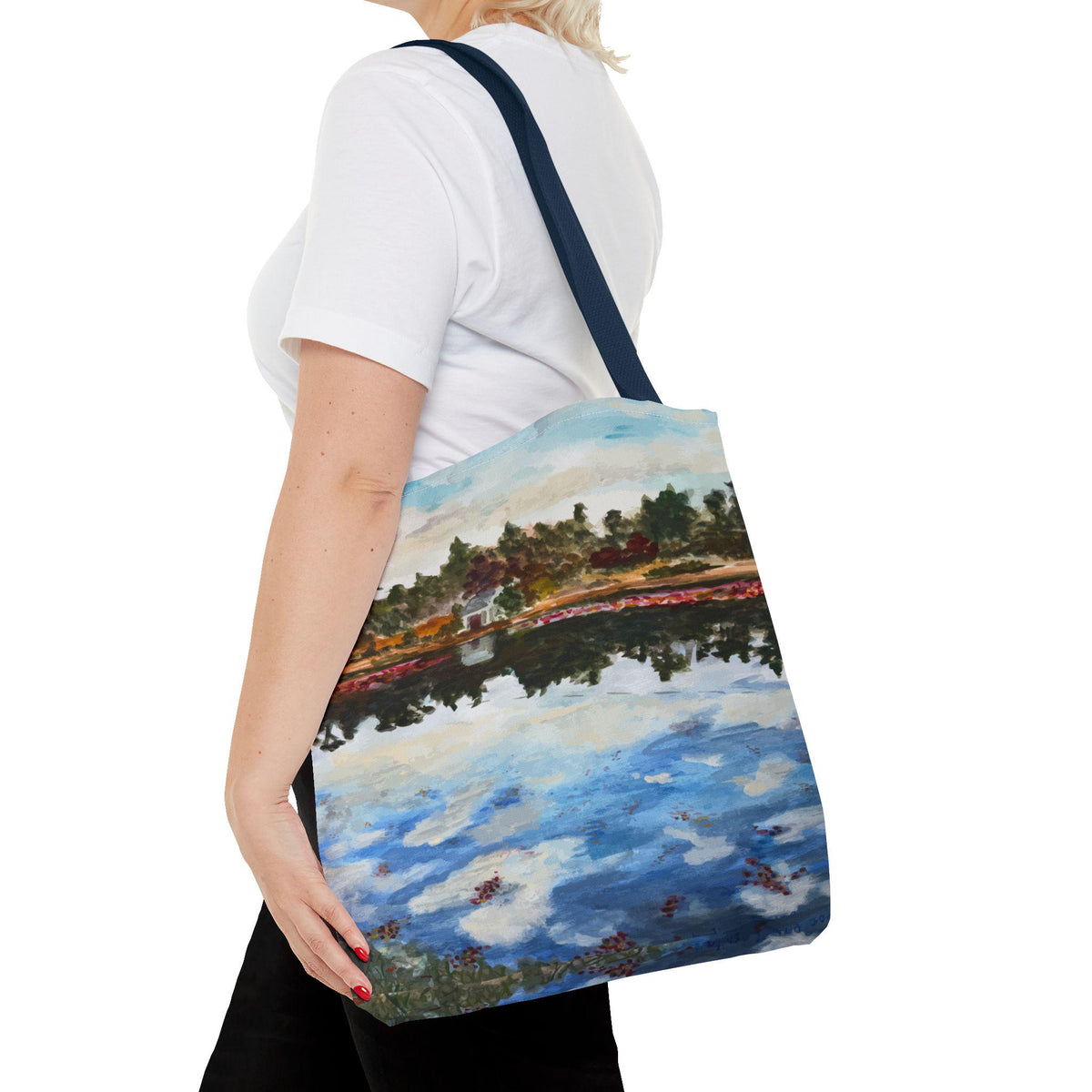 Tote bag with a landscape painting featuring a body of water reflecting a cloudy sky and trees. The bag has a navy blue strap and a white background. The artwork depicts a house and foliage.