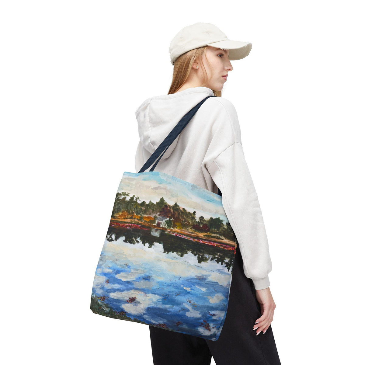 A tote bag with a landscape painting of a lake and trees. The bag has a navy blue strap and is carried over the shoulder. The painting features blue water, green trees, and a white building.