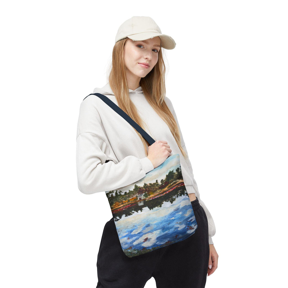 A tote bag with a landscape painting of a lake and trees. The painting features blue, green, brown, and white colors. The bag has a navy blue strap and is being held over the shoulder.