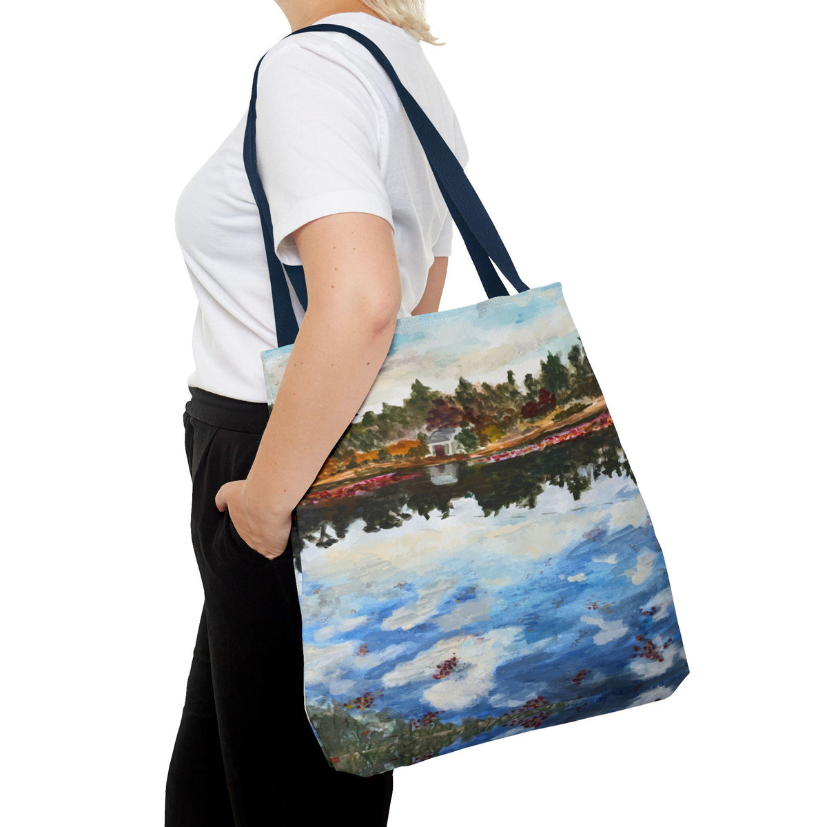 A tote bag with a landscape painting design. The painting depicts a body of water reflecting a shoreline with trees and a building. The bag has navy blue straps and is carried over the shoulder.