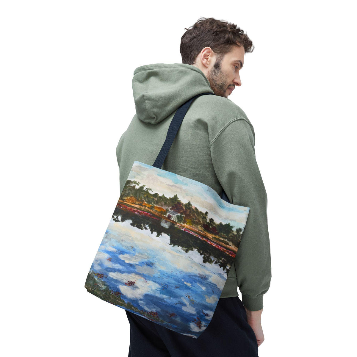 A tote bag featuring a landscape painting with a blue sky, water, and trees. The bag has a navy blue strap and is carried over the shoulder. The painting has a white building in the center.