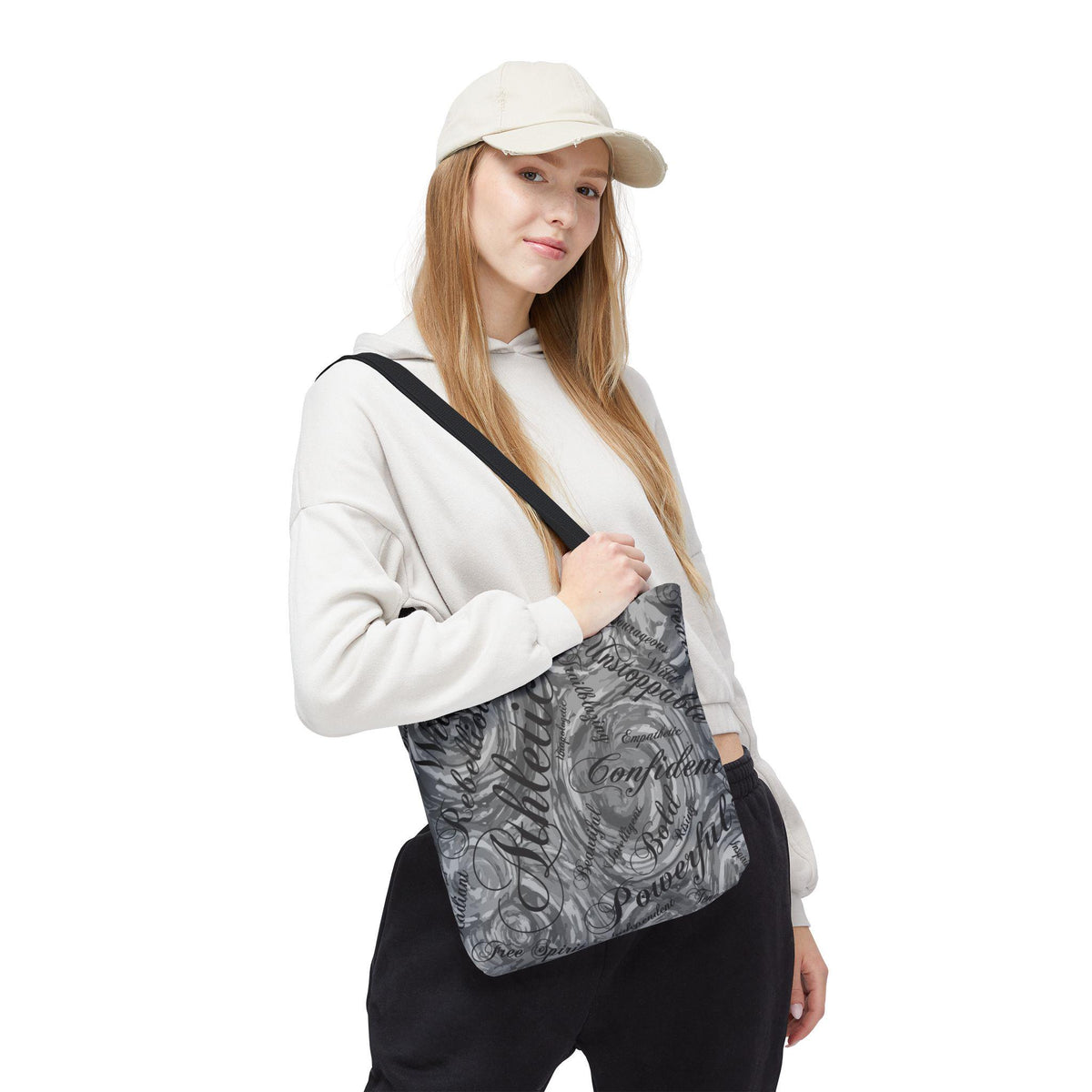 A gray tote bag with a black strap featuring a swirling design and motivational words like "Athlete", "Confident", and "Unstoppable". The bag is held by a person wearing a white hoodie and a cream-colored baseball cap.