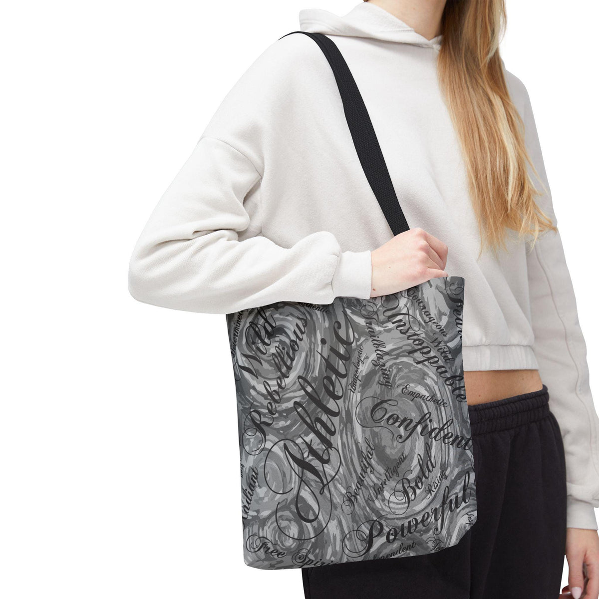 A black tote bag with a swirling gray and white pattern. The bag features words like "Athletic," "Confident," and "Powerful" in a mix of script and block lettering. The bag has a black shoulder strap.