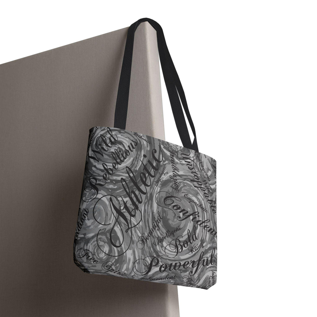 A gray tote bag with black handles, featuring a swirling pattern and words like "Athletic," "Confident," and "Powerful" in a stylish font. The bag is hanging against a neutral-colored wall.