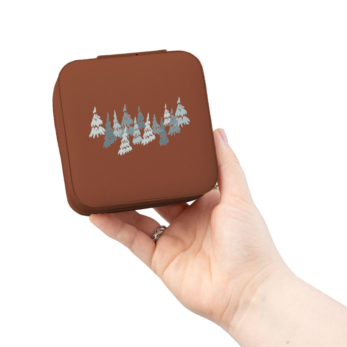 A square, brown trinket box with a forest of stylized evergreen trees in shades of blue and white. The box is held in a hand, showcasing its compact size and design. The box has a hinged lid.