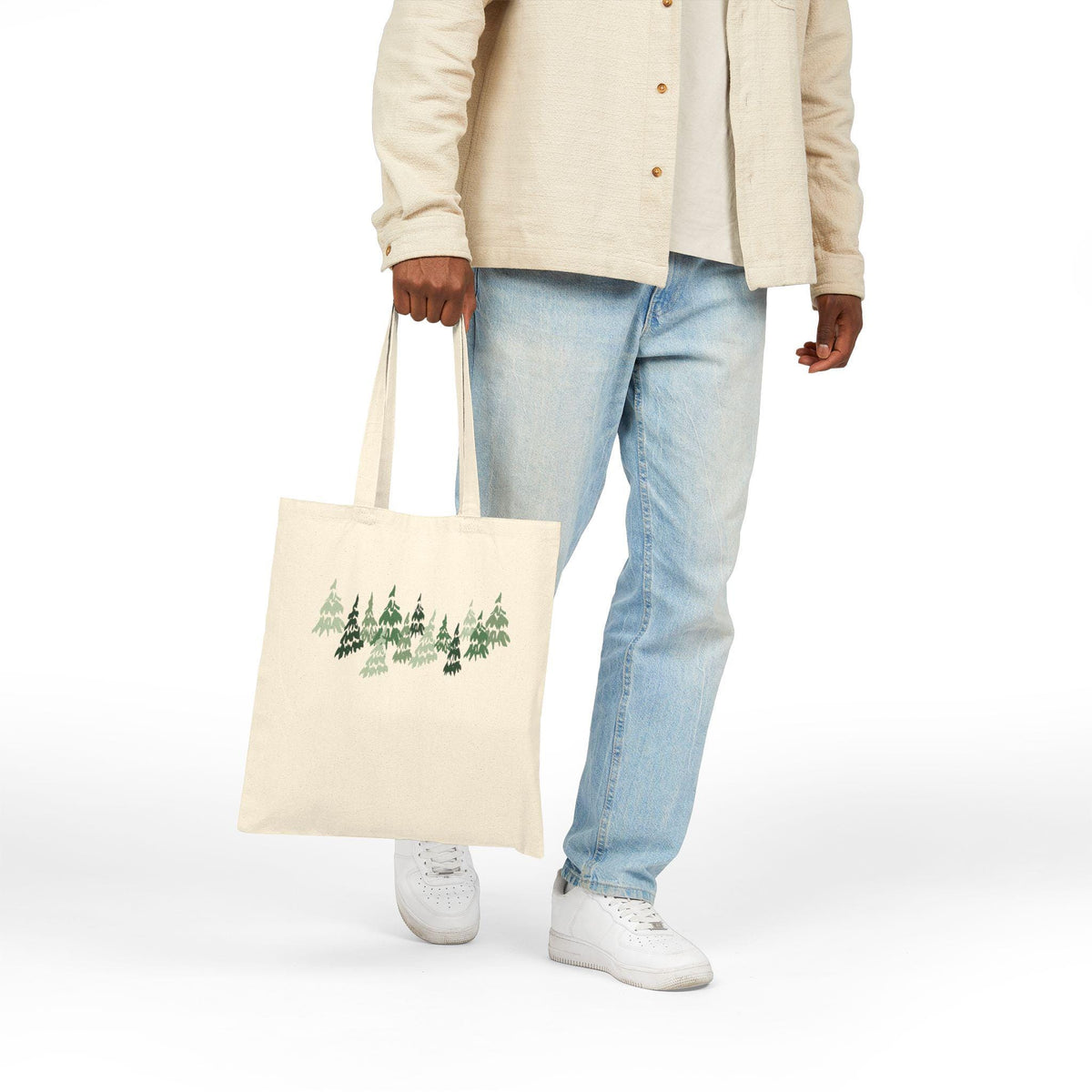 A cream-colored tote bag featuring a forest of green and dark green pine trees. The bag is held by a person wearing a light-colored jacket and blue jeans. The tote bag has long straps.