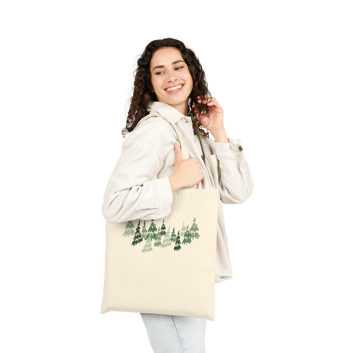 A cream-colored tote bag featuring a forest design in shades of green. The bag is held by a person. The tote bag is made of a sturdy material and is suitable for carrying groceries, books, or other items.