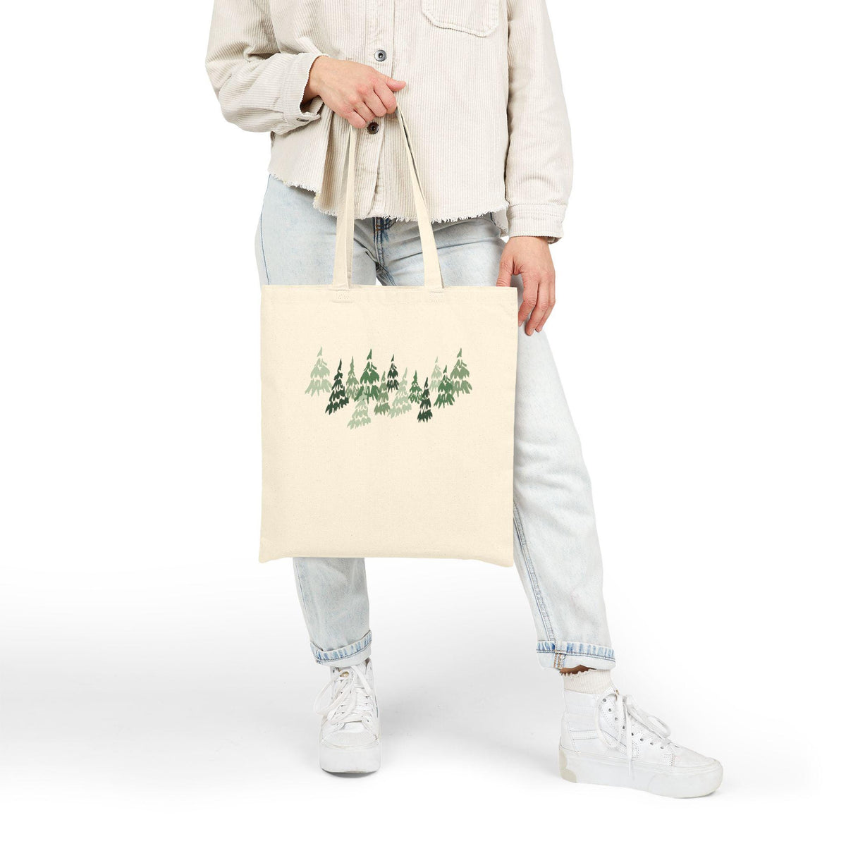 Cream-colored tote bag featuring a forest of green trees. The trees are depicted in varying shades of green, creating a natural and outdoorsy design. The bag is held by a person wearing light-wash jeans and a cream-colored jacket.