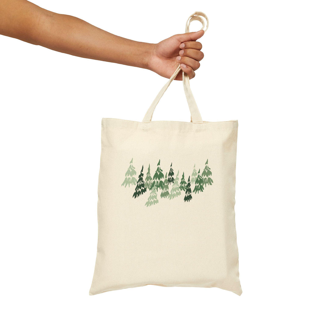 A cream-colored tote bag with a forest design in shades of green. The bag is held by a hand and features a simple, minimalist design. The tote bag is made of a sturdy fabric and has long handles.