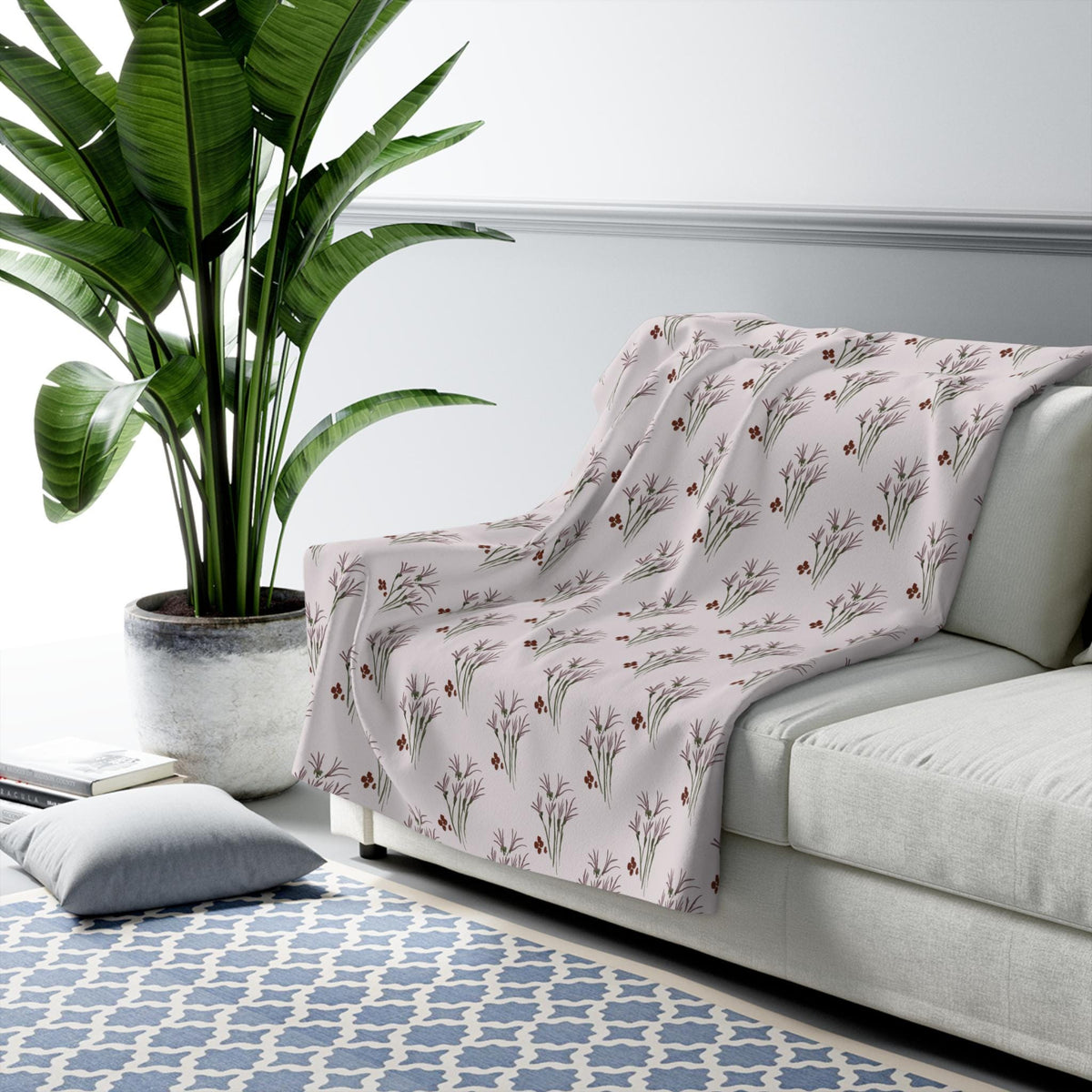 A light pink throw blanket draped over a white sofa. The blanket features a repeating pattern of stylized floral sprigs in shades of brown and green. The sofa is in a room with a large potted plant and a blue patterned rug.