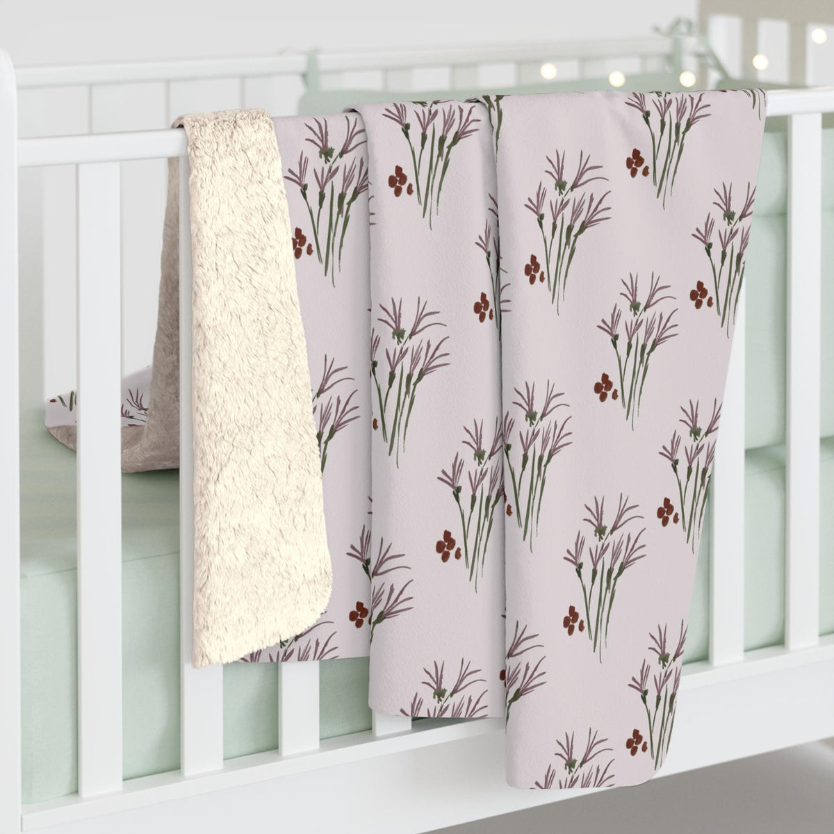 A baby blanket with a floral pattern in shades of purple, green, and red on a light pink background. The blanket is draped over a white crib rail, with a cream-colored sherpa lining visible on the reverse side.