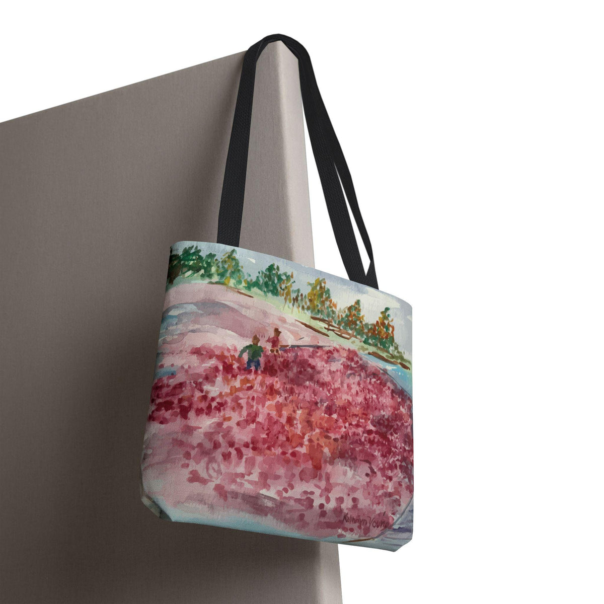 A tote bag with a watercolor painting of a landscape scene. The artwork depicts a field of red flowers, green trees, and a blue sky. The bag has black handles and is hanging against a neutral-colored wall.