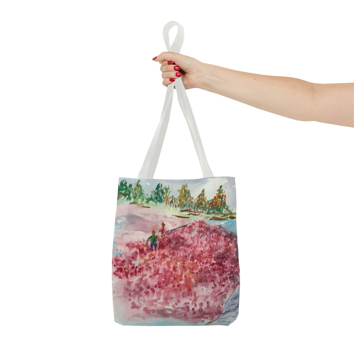 A white tote bag with a watercolor painting of a landscape. The artwork depicts a field of red flowers, green trees, and a blue sky. The bag is held by a person.