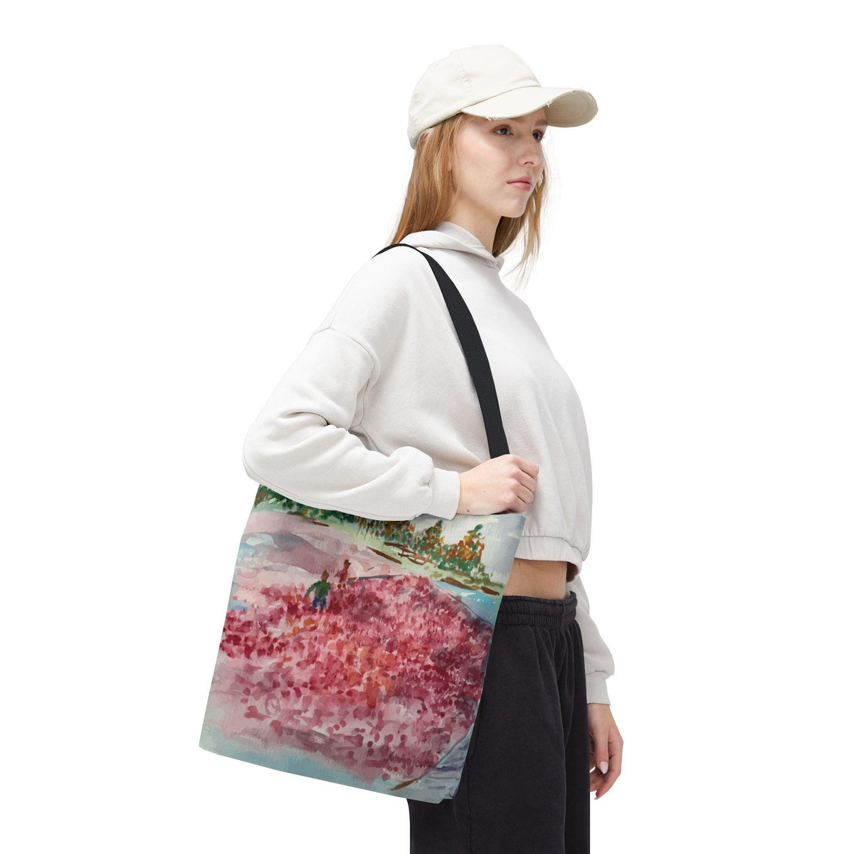 A tote bag with a watercolor painting of a pink floral landscape. The bag has a black strap and is carried over the shoulder. The painting features a field of pink flowers with green trees and a blue sky.