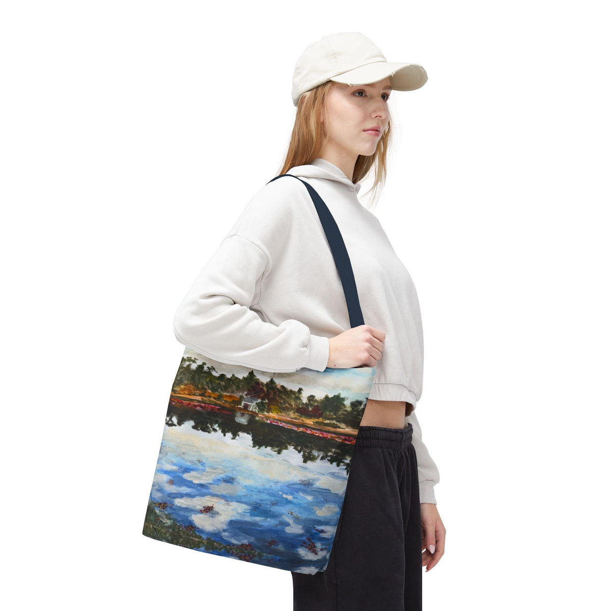A tote bag featuring a landscape painting of a body of water with reflections of the sky and trees. The bag has a navy blue strap and is being carried by a person wearing a white hoodie and black pants.