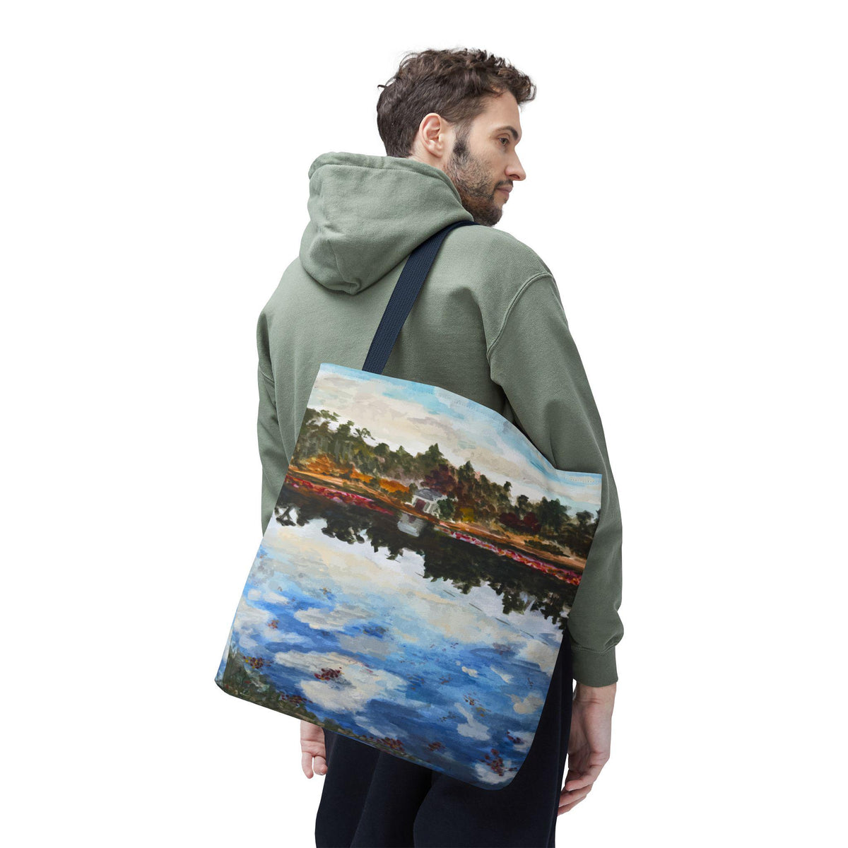 A tote bag featuring an impressionistic painting of a landscape with a lake and trees. The artwork includes blue, white, red, and green hues. The bag has a navy blue strap and is carried over the shoulder.